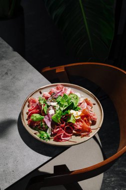 Güzel bir tabak taze yeşillikler ve çeşitli pişmiş etler ile doludur, şık bir masa üzerine kurulur. Loş ışıklandırma rengarenk malzemeleri vurgular, iştah açıcı bir görüntü yaratır.
