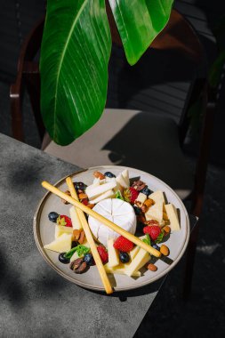 Çok güzel düzenlenmiş bir peynir tabağı, çeşitli peynirler, taze çilekler, yabanmersini, badem ve dekoratif ekmek çubukları içeren bir masaya oturur. Büyük yeşil bir yaprak tropikal bir dokunuş ekler..