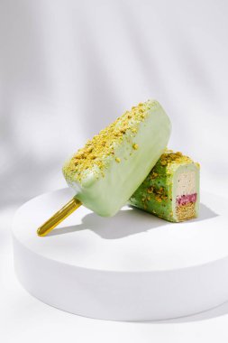 Yeşil dış katmanlı ve pembe merkezli iki buzlu şeker beyaz bir kaidede oturur. Ezilmiş şam fıstıkları tepeyi süslüyor ve sıcak günler için mükemmel bir görsel cazibe yaratıyor..