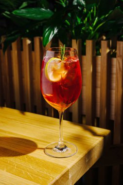 Serinletici bir kokteyl ahşap bir masada oturur. İçecek canlı kırmızı bir renge sahiptir ve limon ve otlarla süslenir. Yeşil bitkiler güneşli ışıkta canlı bir zemin sağlar.