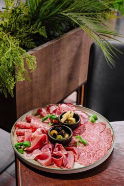 Yuvarlak bir ahşap tabakta, yeşil bir zemin üzerine kurulmuş, siyah zeytin ve şifalı otlarla bezenmiş renkli bir dizi et sergilenir..
