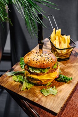 Peynir ve taze sebzelerle dolu doyurucu bir hamburger ahşap bir tahtanın üzerinde duruyor. Küçük bir patates kızartması sepeti şık bir restoranda hazırlanan yemeğe eşlik ediyor..