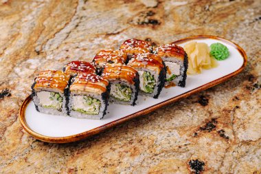 Taze hazırlanmış suşi ruloları beyaz bir tabakta sergilenir. Ekmekler parlak bir üst kaplama içeriyor ve onlara parlak yeşil wasabi ve mermer bir yüzey üzerinde turşulu zencefil eşlik ediyor..