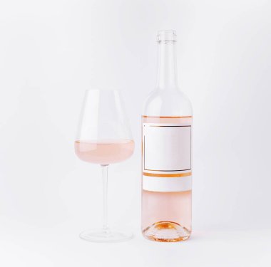 Açık pembe gül şarabıyla dolu berrak bir bardak beyaz arka planda bir gül şişesinin yanında oturur. Şişedeki etiket boş, içkinin canlandırıcı görünümünü vurguluyor..