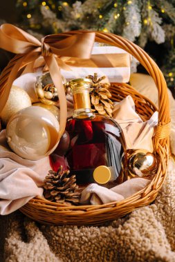 Dekoratif bir şişe, altın süsler ve kozalaklarla dolu bir sepet rahat bir ortamda oturur. Yumuşak kumaş ve paketlenmiş hediyeler bayram cazibesine katar.