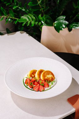 Canlı taze çileklerle kaplı bir tabak altın krep zarif, modern bir masada servis edilir. Yeşil bitkiler arka planda taze bir atmosfer yaratarak yemek deneyimini güçlendirir..