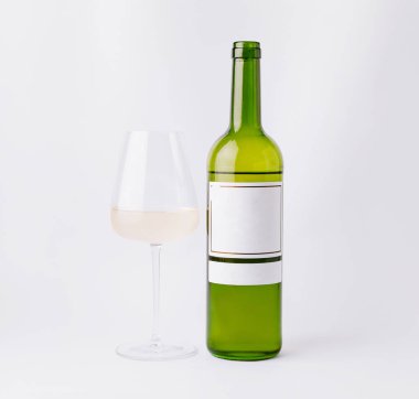 Beyaz şarapla dolu yeşil bir cam şişe berrak bir şarap kadehinin yanında duruyor. Şarabın zarafetini vurgulayan, sade beyaz bir arka planı olan, minimalist bir ortam..