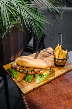 Marul, domates ve diğer malzemelerle birlikte taze yapılmış bir sandviç ahşap bir tahtanın üzerinde duruyor. Yanında küçük bir patates kızartması sepeti var. Hepsi de dinlendirici bir iç mekanda sunuluyor..