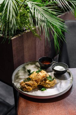 Kızarmış tavuk parçaları, içinde sos bulunan iki küçük kâse ile şık bir tabakta sunulur. Yeşil bir bitki arka planı dekore ederek modern bir restoran ortamı yaratıyor..