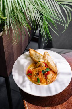 Bir kase lezzetli deniz ürünleri güveci, yanında ekmek dilimleriyle birlikte ahşap bir masada oturur. Yeşil bitkiler sıcak atmosfere tazelik katar, sıcak bir yemekten zevk almak için mükemmeldir..