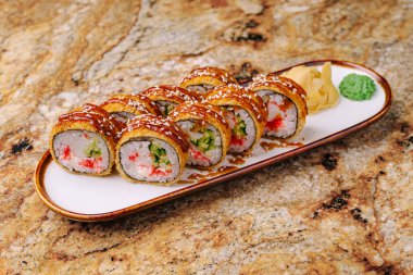 Suşi yuvarlakları, desenli granit bir yüzeyde beyaz bir tabakta düzgünce dizilmiş. Ekmekler sosla kaplanmış, susam, zencefil ve wasabi ile süslenmiş..