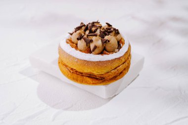 Taze yapılmış bir tatlı beyaz bir tabakta oturur. Sergide kremsi dolgu maddeleri ve çikolata talaşlarıyla kaplı yumuşak hamur işleri yer alıyor. Sunum onun tatlı tasarımını vurgular.