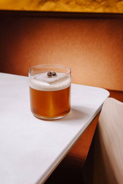 Bir kahve kokteyli parlak beyaz bir masanın üzerindeki bir bardakta zarif bir şekilde durur. Yerleşim, sıcak topraksı tonlara sahip, içeceklerden keyif almak için rahat bir atmosfer yaratıyor..