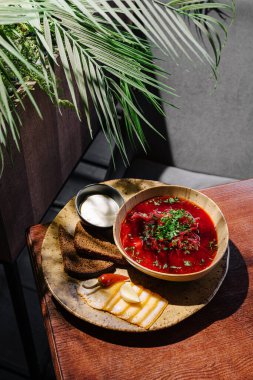 Renkli bir kase pancar çorbası, yanında ekmek ve peynir dilimleri ile birlikte ahşap bir masada oturur. Taze otlar çorbayı süslüyor, iştah açıcı sunumu güçlendiriyor..