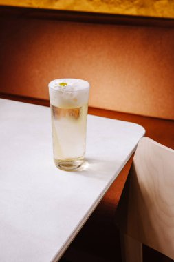 Temiz bir içecekle dolu uzun bir bardak beyaz bir masada duruyor. İçkinin üzerinde köpük ve küçük bir çiçek var. Sıcak ışık altında davetkar bir atmosfer yaratıyor..