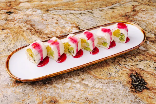 Çok güzel hazırlanmış bir suşi tabağında balık ve sebze katmanlı birden fazla rulo var. Tabak renkli bir sos ile zenginleştiriliyor ve parlak beyaz bir tabakta sunuluyor..