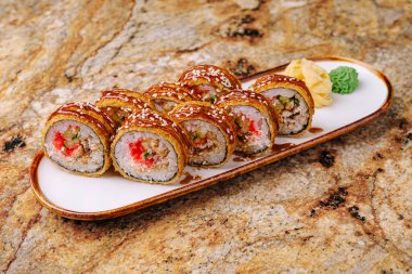 Lezzetli suşi ruloları, wasabi ve zencefil turşusuyla süslenmiş uzun beyaz bir tabakta sunuluyor. Bu rulolarda rengarenk malzemeler ve kıtır kıtır bir doku var. Deniz ürünleri severler için mükemmel..