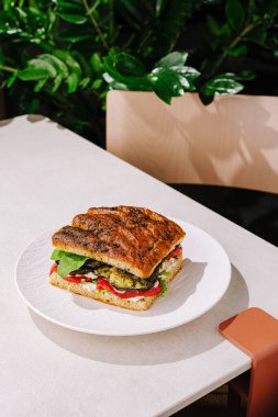 Taze sebzelerle dolu ağız sulandıran bir sandviç beyaz bir tabakta oturur. Yeşillik bitkilerle çevrili bir kafede basit bir masada sunuluyor, rahat bir atmosfer yaratıyor..