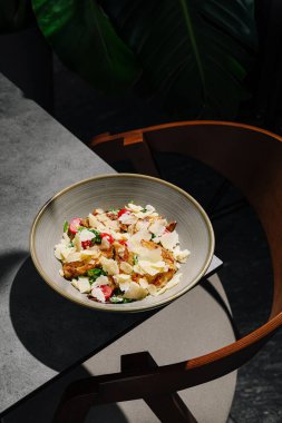 Taze yeşillik, turp ve sosla dolu renkli bir salata masada bir kasede duruyor. Yakınlarda, şık bir sandalye modern yemek düzenini tamamlayarak canlı bir atmosfer yaratıyor..