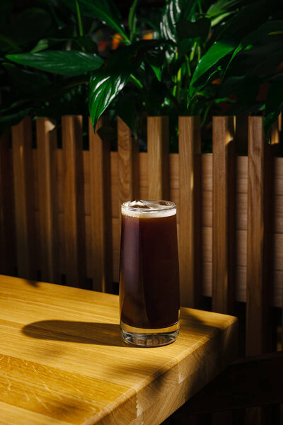 A tall glass filled with a dark, refreshing beverage is placed on a wooden table. Lush green plants in the background enhance the warm, inviting atmosphere.