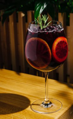 Renkli bir kokteyl ahşap bir masanın üzerinde zarif bir şekilde duruyor. Kan portakalı dilimleri ve yeşil bir bitki filizi sergiliyor. İçkiler gün ışığında parlıyor, ferahlatıcı bir deneyime davetiye çıkarıyor..