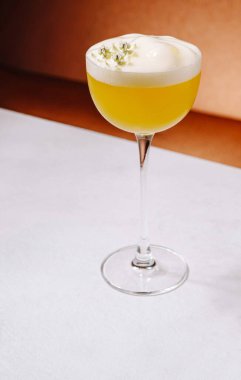 Parlak sarı bir karışım zarif bir bardakta duruyor köpüklü bir sosla süslenmiş ve küçük otlarla süslenmiş. İçki yumuşak bir zemine yerleştirilir, sıcak bir zemine karşı..