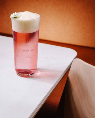 Köpüklü pembe bir içecekle dolu uzun bir bardak ve beyaz bir masada küçük bir çiçek oturuyor. Arka plan, içkiyi tamamlayan sıcak ve dünyevi tonlara sahiptir..