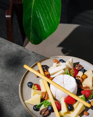 Çok güzel düzenlenmiş bir peynir tabağında çeşitli peynirler, taze çilekler, yabanmersini ve fındık bulunur. Güneş ışığı sahneyi nazikçe aydınlatır, renkleri ve dokuları güçlendirir.