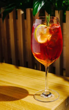 Canlı bir kokteyl, limon ve biberiyeyle süslenmiş ahşap bir masada oturur. Sıcak atmosfer ve yeşillik arkadaşlarla içki içmek için rahatlatıcı bir ortam yaratır..