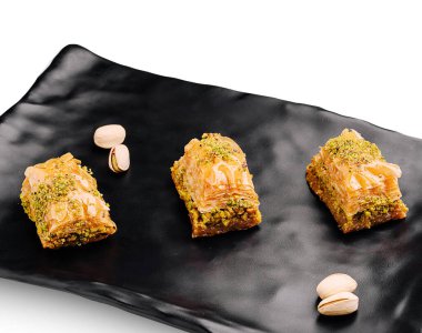 Baklava, altın katmanları ve narin şam fıstığı kaplamasını gösteren siyah bir tabakta zarif bir şekilde düzenlenmiştir. Zengin tatlıya katmak için saçılmış fıstıklı fındıklar eşlik eder..