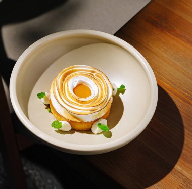 Lezzetli tatlı üzerine kremalı girdaplar ve nane yapraklarıyla süslenmiş yuvarlak bir hamur işi içerir. Basit bir kasede servis edilen bu ikram zarif ve davetkar görünüyor..