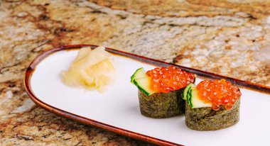 İki suşi rulosu, canlı balık yumurtası ve salatalıkla kaplı, parlak bir tabakta oturur. Zencefil turşusu bir tutam tat katar. Ayarda desenli taş bir yüzey yer alıyor..