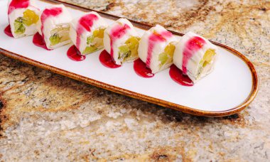 Uzun bir tabakta düzgün sıralanmış tatlı suşisi canlı tatlı meyve ve krema katmanları içerir. Altına serpiştirilmiş sos bu eşsiz yemeğe sanatsal bir dokunuş katıyor..