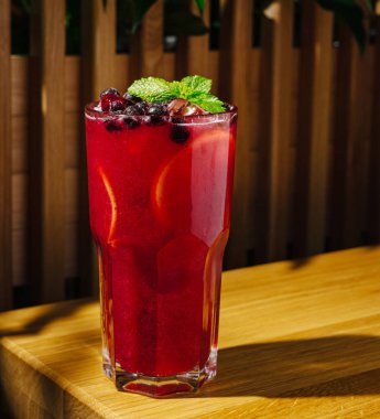 Canlı kırmızı böğürtlen içeceğiyle dolu uzun bir bardak ahşap bir yüzeye oturur. İçeceğe taze nane ve narenciye katılmış..