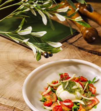 İçinde mozzarella burrata, kiraz domatesleri ve taze otlar bulunan lezzetli bir salata kırsal ahşap bir masada sergilenmektedir. Zeytin dalları olay yerine taze bir dokunuş sağlıyor..
