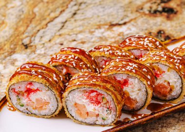 Bir tabak çıtır suşi rulosu, zengin bir sos ve dağınık susam tohumlarıyla kaplı deniz ürünleri ve krema karışımı içererek lezzetli bir mutfak deneyimi yaratıyor..