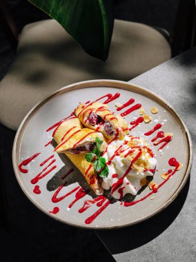 Bir tabakta taze meyve ve kremayla süslenmiş, üzerine kırmızı sos serpiştirilmiş iki krep var. Yumuşak doğal ışık, rahat bir kafe ortamındaki renkli tatlıyı ön plana çıkarıyor..