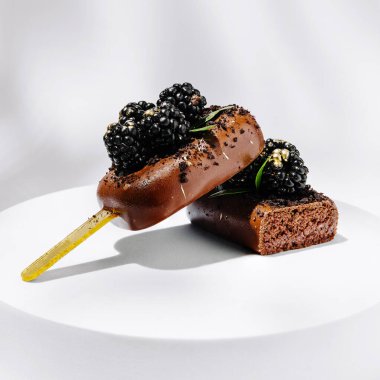 Lezzetli bir tatlı, üzerinde beyaz bir tabakta cezbedici bir ikram sunan taze böğürtlenlerle kaplı kare bir kekin üzerinde dinlenen çikolatalı dondurmayı içerir..