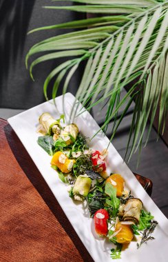 Sarılmış sebzeler, taze sebzeler ve otlar içeren canlı bir yemek uzun beyaz bir tabakta zarif bir şekilde düzenlenmiştir. Yumuşak gölgeler yemeğin renklerini ve dokusunu vurgular.