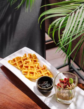 Bir bardak kremalı dondurma ve zengin sosla birlikte nefis bir tabak altın waffle 'ın tadını çıkarın. Sahne yemyeşil yapraklarla dolu, canlı bir atmosfer yaratıyor..