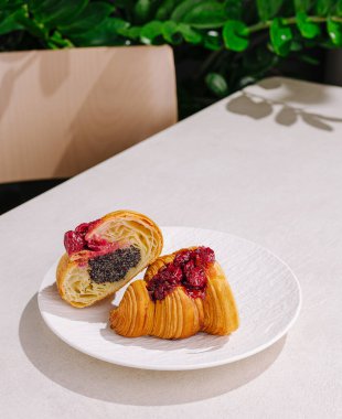 İki yeni pişmiş kruvasan beyaz bir tabakta sergilenir. Birinde zengin haşhaş tohumu dolgusu, diğerinde ise yeşil bir zemin üzerine kurulmuş canlı ahududu kompostosu var..