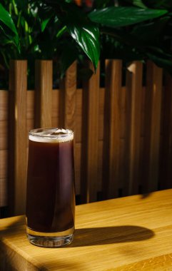 Büyük bir bardak soğuk kahve, kapalı bir alanda ahşap bir masada oturur. Arkaplanda yemyeşil bitkiler var. Sıcak ve davetkar bir atmosfer yaratıyorlar. Rahatlamak için mükemmel..