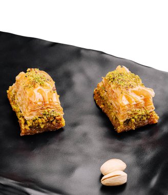 Üzerinde ezilmiş şam fıstığı olan iki parça tatlı baklava siyah bir tabakta duruyor. Tatlının altın bir tabakası ve parlak bir kaplaması var. Zengin, fındık dolgulu dokusunu sergiliyor..