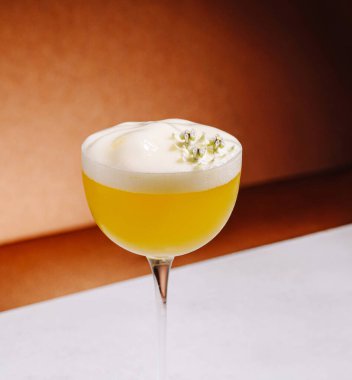 Canlı sarı bir kokteyl köpüklü bir üst ile zarif bir bardakta oturur. Küçük çiçekler içeceği süslüyor, sıcak bir arka plana biraz zarafet katıyor..