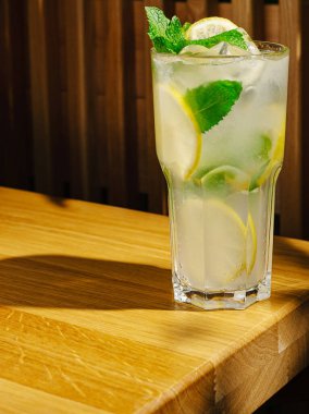 Taze nane yaprakları ve limon dilimleri ile dolu uzun bir bardak mojito pürüzsüz bir ahşap masada oturur. Güneş ışığı içkiyi vurgular, rahatlamak için mükemmel bir karşılama atmosferi yaratır..