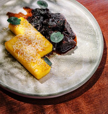 Canlı bir tabak, zengin, koyu bir sosun yanında peynirle pişirilmiş polenta içerir. Taze otlar bir parça renk katar, bir restoranda gurme mutfağı sergiler..