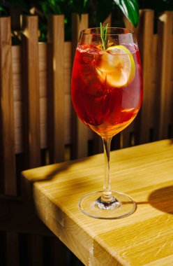 Canlı kırmızı bir içecekle dolu uzun bir bardak ahşap bir masada oturur. Taze meyve ve bir tutam biberiye içeceği süslüyor. Yumuşak ışık sıcak bir atmosfer yaratır..