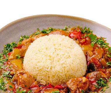 Bir kase renkli sebzeli tavuk güvecinin üzerinde yumuşacık bir pirinç yığını içerir. Tabak taze otlarla süslenmiş, bu da onu cazip bir yemek seçeneği yapıyor..