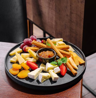 Lezzetli peynir ve meyve tabağında çeşitli peynirler, taze üzümler, kurutulmuş meyveler ve küçük bir kase reçel yer alıyor. Ortam sıcak ve davetkar, paylaşmak için mükemmel..