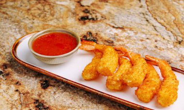 Lezzetli karides, altın ve çıtır çıtır olana kadar mükemmel bir şekilde kızartılır, uzun bir tabakta güzelce dizilene kadar. Küçük bir kase tatlı sos yanında oturur ve lezzetli bir deneyimi davet eder..